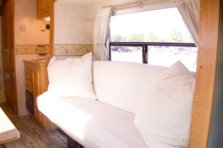 Bright RV rental interior featuring a white sofa with pillows, a small kitchenette, and a window overlooking a parking lot.. R-Vision Trail-Lite 2002