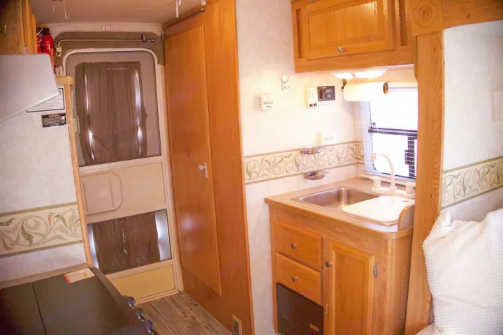 Cozy RV rental interior showing a compact kitchen with a sink, wooden cabinets, and the main entrance door for travel.. R-Vision Trail-Lite 2002