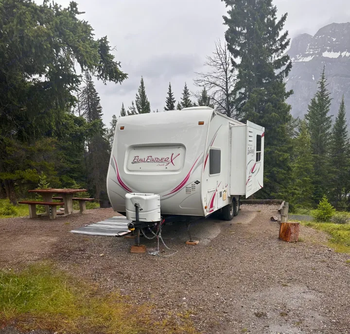 This rv rental, a white travel trailer, is nestled in a gravel campsite surrounded by pine trees and distant mountains, ideal for camping.. Cruiser RV Fun Finder X 2011