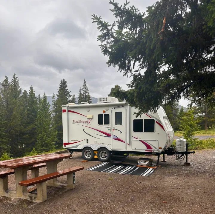This RV rental is set up at a scenic campsite, featuring a white travel trailer, picnic table, and evergreen trees under an overcast sky.. Cruiser RV Fun Finder X 2011