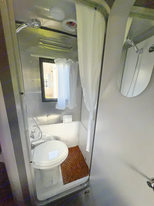 Inside an RV rental bathroom: compact shower/toilet combo, window with curtains, and wooden slat floor.. Airstream Basecamp 2025