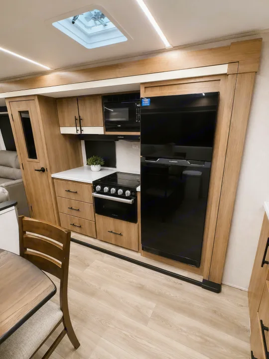 Modern RV rental kitchen featuring light wood cabinetry, black appliances, white counters, and dining area.. Grand Design Imagine 2024