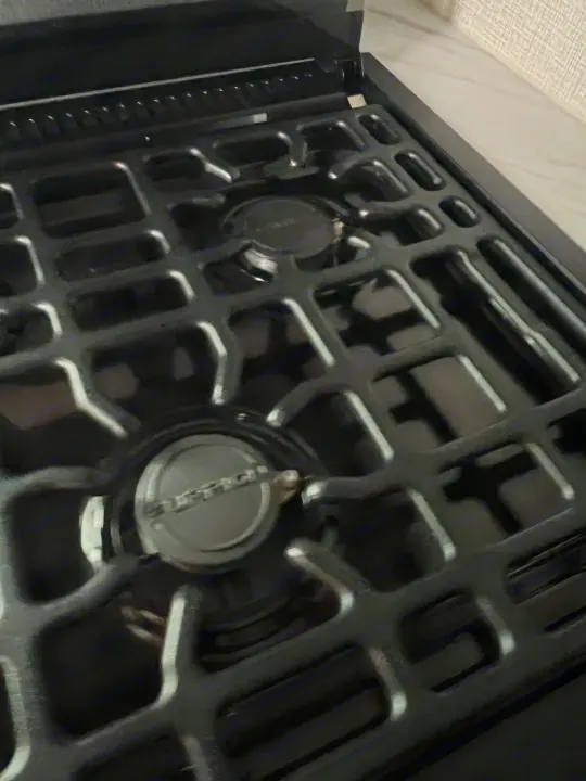 RV rental kitchen: Close-up of a dark, glossy gas stove with two burners and sturdy grates, ideal for preparing meals on your trip.. Grand Design Imagine XLS 2024