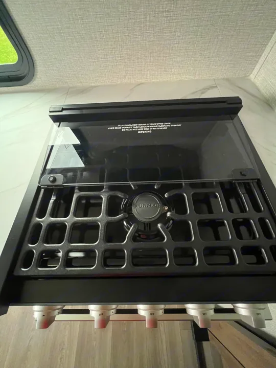 Close-up of a modern Furrion rv rental cooktop with open glass cover, burner grates, and controls, ready for cooking.. Grand Design Imagine XLS 2024