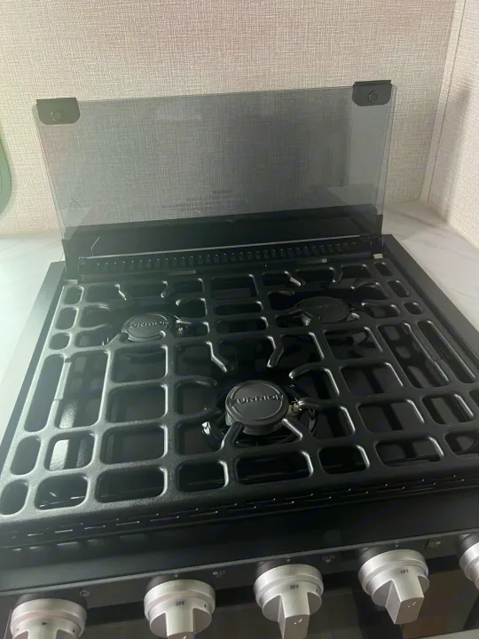 A clean Furrion gas cooktop with three burners and a raised glass cover, ideal for an RV rental kitchen. Warning visible.. Grand Design Imagine XLS 2024
