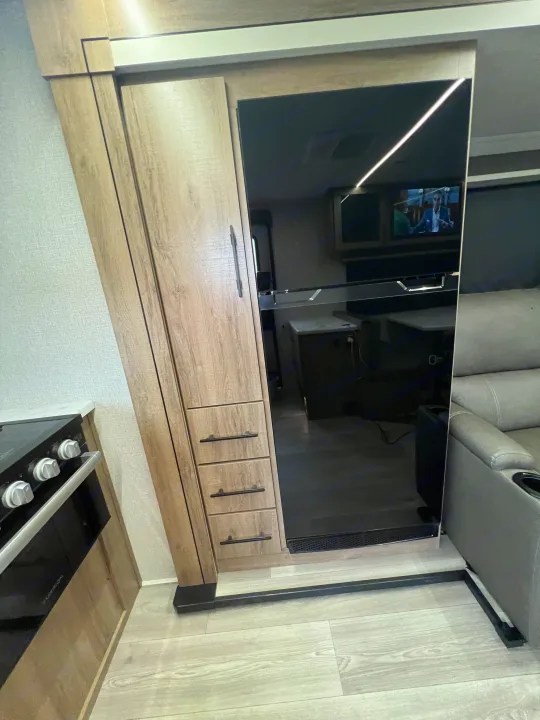 Modern RV rental interior with a wood-grain cabinet, mirrored sliding door reflecting the living area, and part of the kitchenette.. Grand Design Imagine XLS 2024