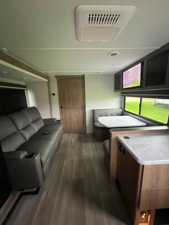 Spacious rv rental interior with a gray sofa, dining booth, kitchen counter, wood floor, and overhead AC unit.. Grand Design Imagine XLS 2024
