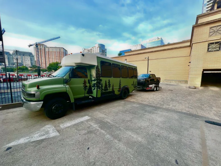 Green RV rental bus with tree decals, towing a black pickup truck on a U-Haul trailer, parked in an urban area.. Chevy C4500 Kodiak 2008