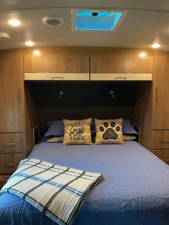 This RV rental features a comfortable bedroom with a blue quilted bed, 'Happy Camper' pillows, overhead storage, and ceiling vent.. Grand Design Imagine 2022