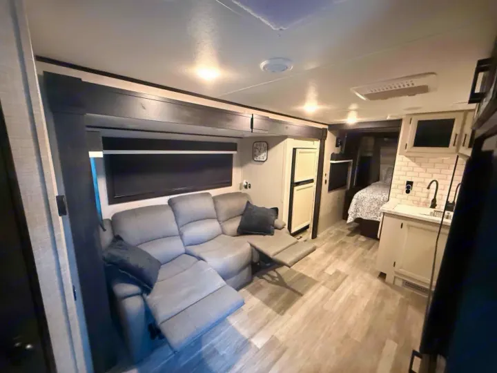 Modern rv rental interior: gray sectional with recliners, full kitchen, and bedroom view. Light wood floors, overhead lighting.. Jayco Jay Feather 2023
