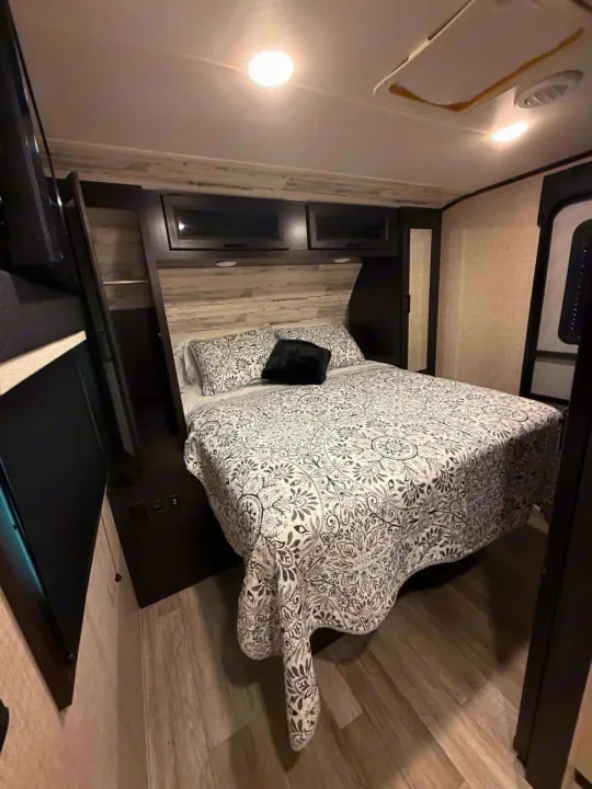 Modern RV rental bedroom with a queen bed, stylish black & white patterned bedding, overhead storage, and wood-look floor.. Jayco Jay Feather 2023