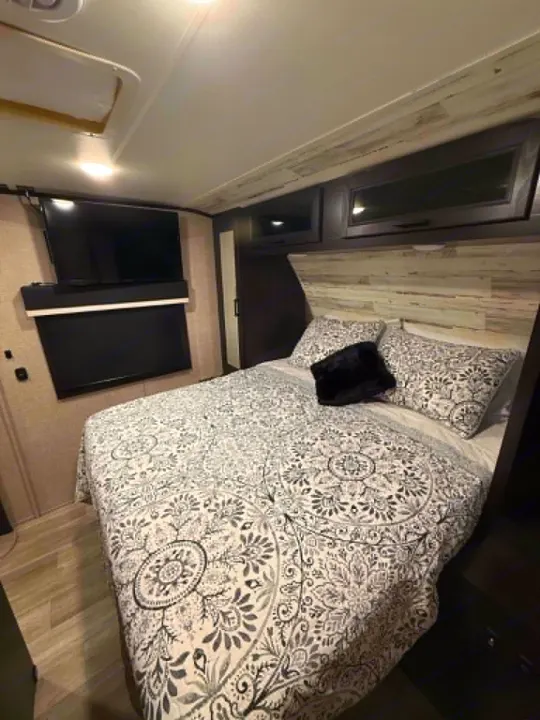 Cozy RV rental bedroom with patterned bed, wall-mounted TV, and overhead storage. Ideal for comfortable travel.. Jayco Jay Feather 2023