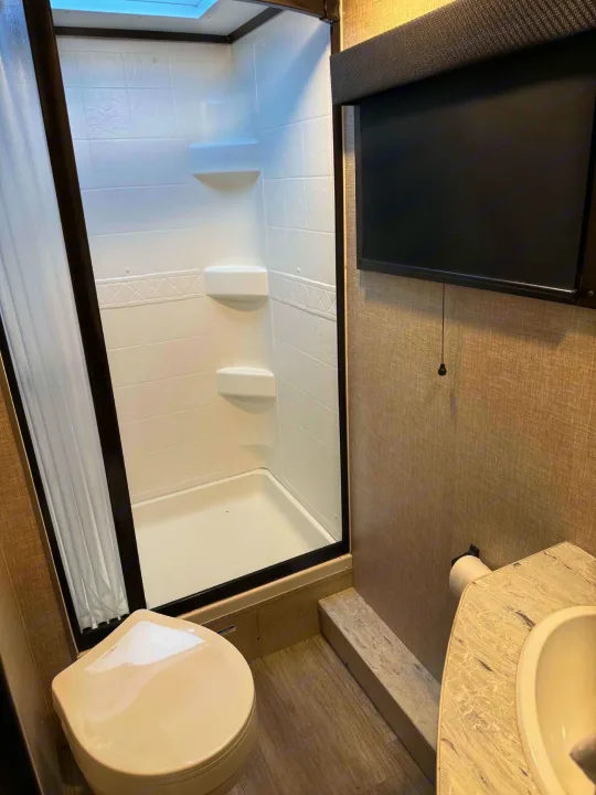 This rv rental bathroom features a clean shower stall, toilet, and sink, with a skylight and window blind for comfort.. Jayco Jay Feather 2023