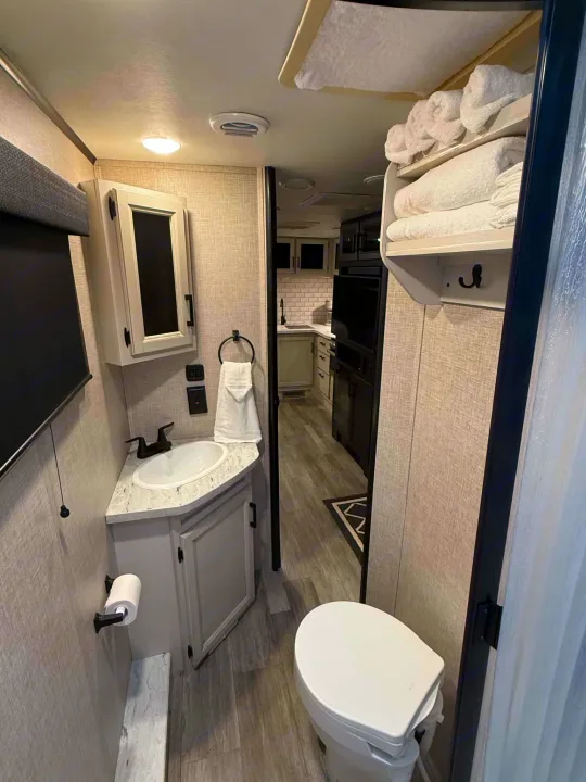 This clean RV rental bathroom offers a toilet, sink, vanity, mirrored cabinet, and shelves stocked with fresh towels.. Jayco Jay Feather 2023