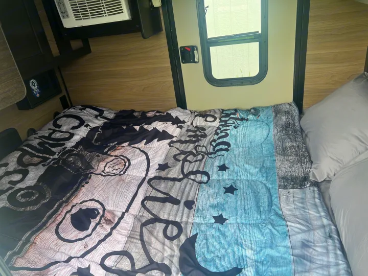 RV rental bedroom features a unique wood-patterned comforter, grey pillow, interior window, and AC unit.. BUSHWACKER 10HD 2024