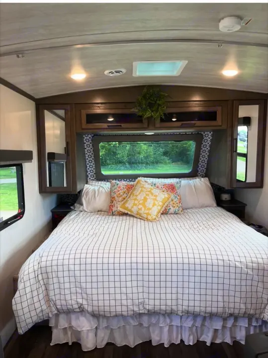 Cozy RV rental bedroom with checkered duvet, floral pillows, overhead storage, and a scenic window view.. Keystone RV Cougar 2023