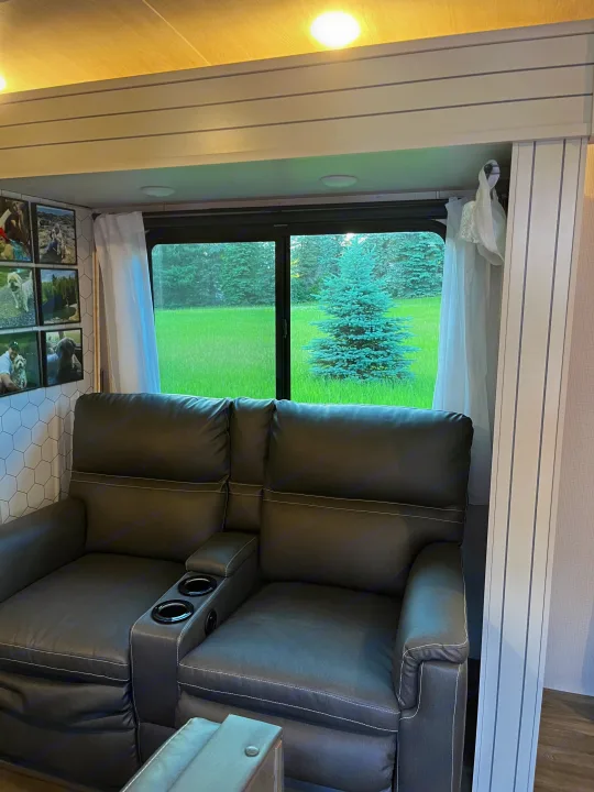 Cozy RV rental living space featuring a dark gray reclining sofa with cup holders and a scenic window view of pine trees.. Keystone RV Cougar 2023