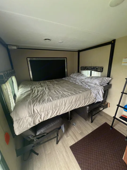 This RV rental interior showcases an elevated bed with light bedding and leaf-patterned pillows, positioned over a seating area, with a dark floor mat.. Coleman Lantern Series 2016