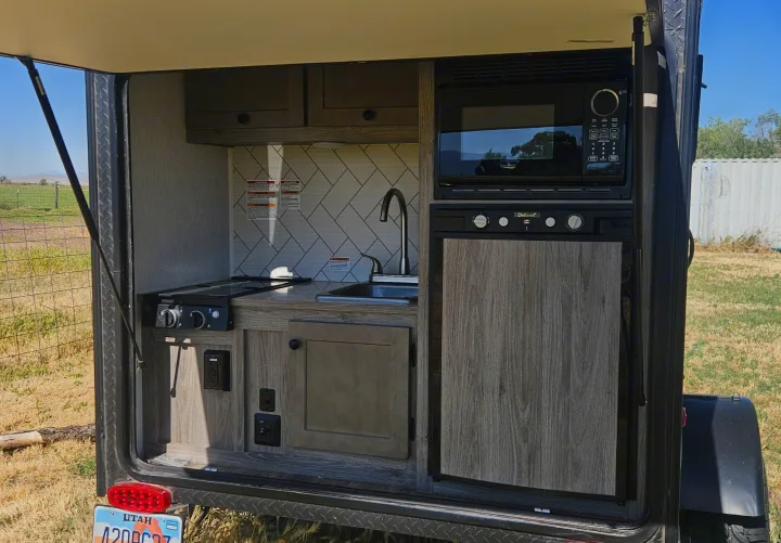 This compact RV rental features a well-equipped outdoor kitchen with a microwave, fridge, sink, stovetop, and cabinets.. Sunset Park RV SunRay 2024