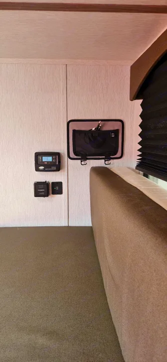 This RV rental interior features a solar controller, Dometic panel, USB port, and fabric storage pouch next to a cushioned bed.. Sunset Park RV SunRay 2024