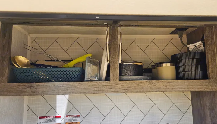 Organized RV rental kitchen cabinet with utensils, pots, cups, an electrical outlet, and safety warnings below.. Sunset Park RV SunRay 2024