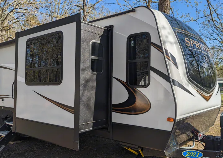 This RV rental, a white travel trailer with an extended slide-out, is parked at a sunny campsite.. Keystone RV Sprinter Limited 2021