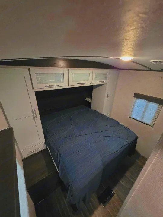 Cozy rv rental bedroom featuring a bed with dark blue sheets, white cabinets, and a window with blinds for comfortable stays.. Keystone RV Bullet Ultra Lite 2021