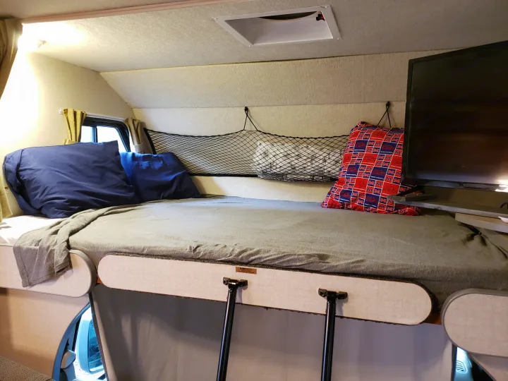 RV rental interior featuring a comfortable cab-over bunk bed, overhead storage net, and TV, perfect for travel.. Fleetwood Tioga 31M 2007