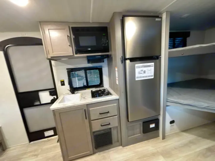 Inside an RV rental: a compact kitchen with microwave, sink, stovetop, and fridge, next to cozy bunk beds. Ideal for travel.. Keystone RV Hideout Sport 2025