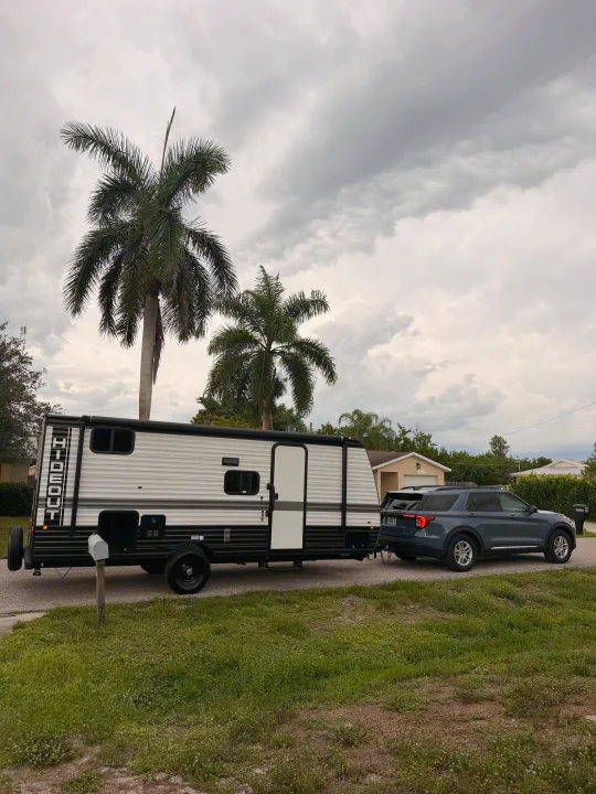RV rental: Dark SUV towing a white 'Hideout' travel trailer on a street under cloudy skies, flanked by palm trees.. Keystone RV Hideout Sport 2025