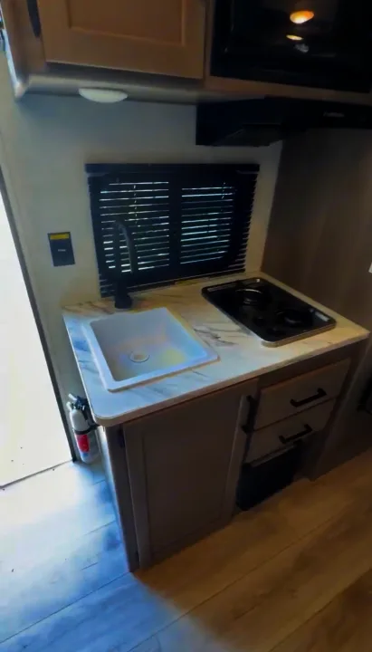 Compact RV rental kitchen with white sink, black faucet, 2-burner stove, microwave, and light gray cabinets.. Keystone RV Hideout Sport 2025