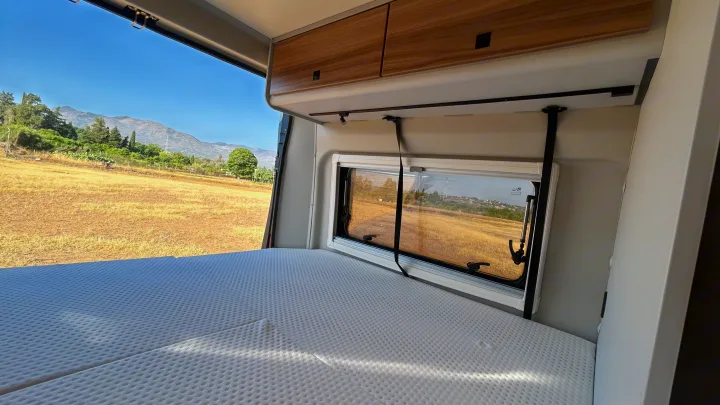 Inside an **RV rental**: a comfortable bed facing a window with a beautiful natural view of dry fields and distant mountains.. BURSTNER B66 2024