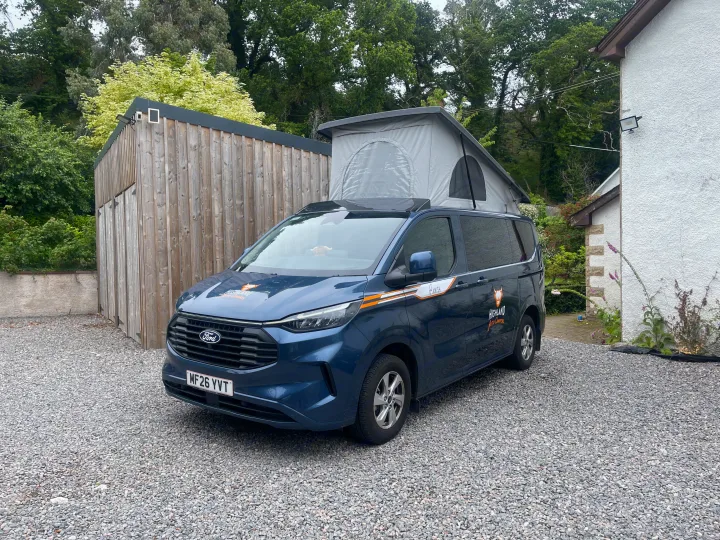 Blue Ford campervan RV rental with pop-up roof, parked on a gravel driveway next to a wooden building and a white house.. Ford Transit Custom 2+2 berth Pop Top, Automatic 2026