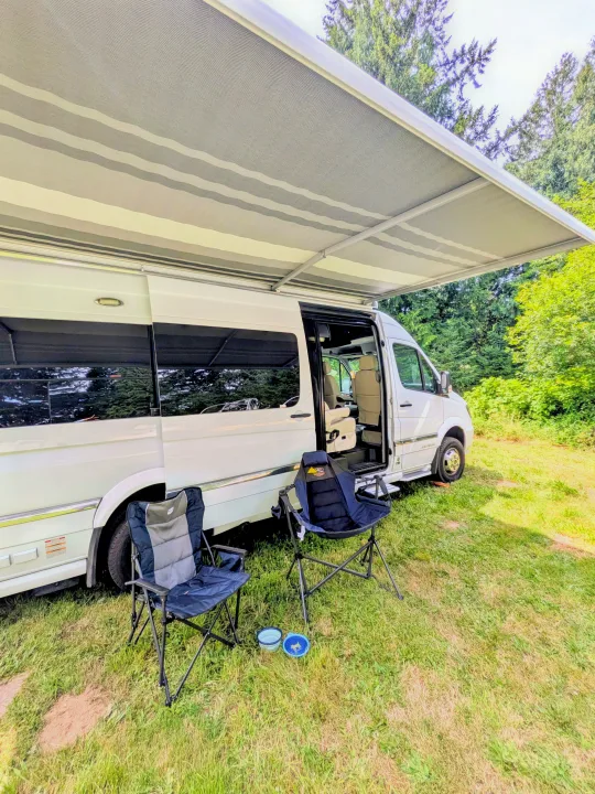 A white RV rental with an extended striped awning, two camping chairs, and pet bowls on the grass, surrounded by trees.. Airstream Interstate 2017