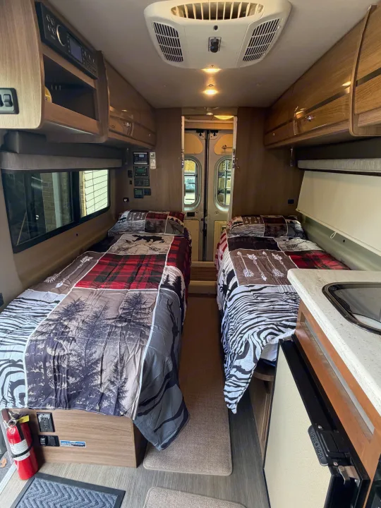 RV rental interior: two single beds with patterned comforters, overhead storage, and a compact kitchen area are visible.. Winnebago Travato 2016