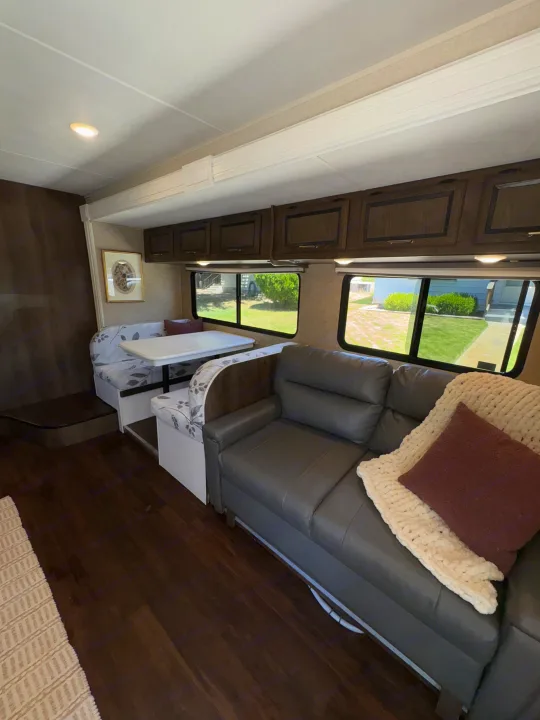 Cozy RV rental interior featuring a gray sofa, dinette with floral cushions, dark wood cabinets, and large windows.. Coachmen Freelander 2018