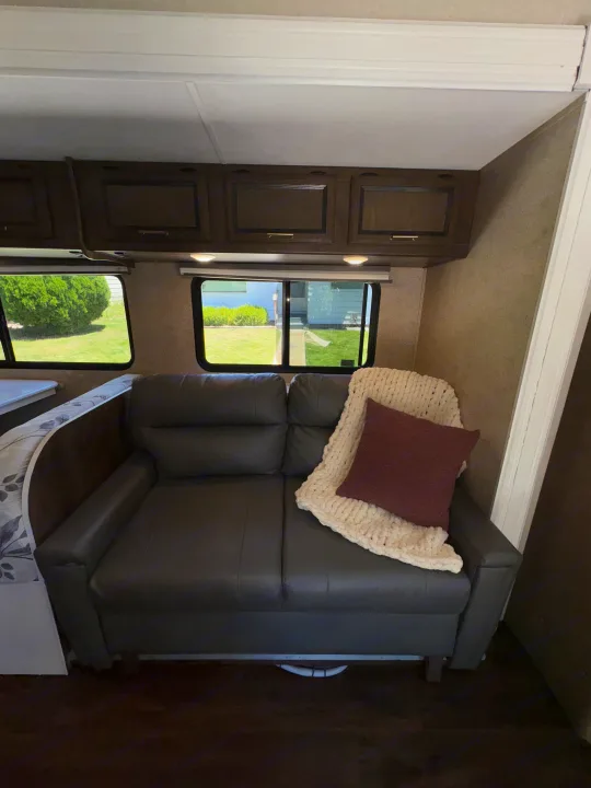 Cozy RV rental living space: dark couch, soft blanket, nature views from windows, and overhead wood cabinets.. Coachmen Freelander 2018