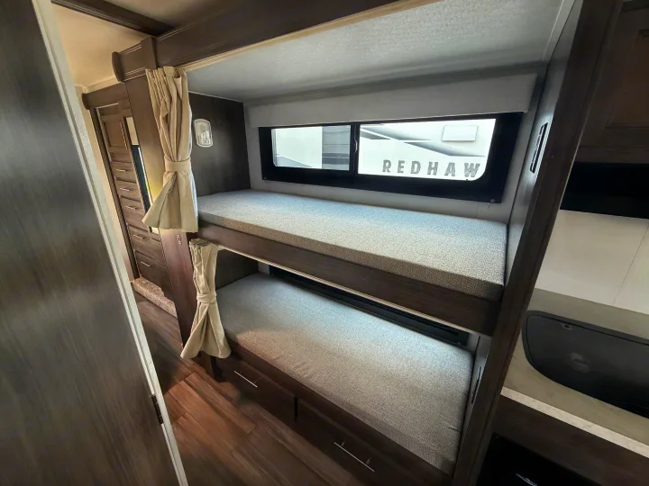 Interior view of an RV rental featuring modern bunk beds with grey cushions and tied-back curtains. A window shows 'REDHAW' branding.. Jayco Redhawk SE 31FF 2026