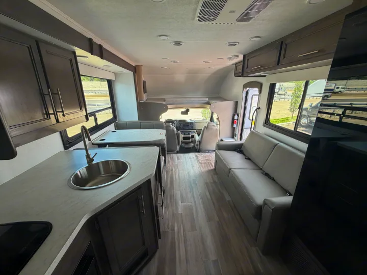Bright, modern rv rental interior with kitchen sink, dinette, sofa, and driver's cabin visible, featuring wood-look floors.. Jayco Redhawk SE 31FF 2026