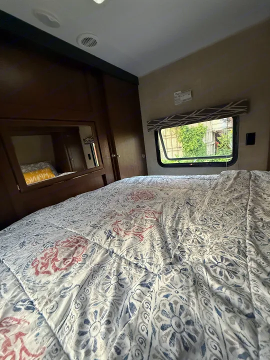 Cozy RV rental bedroom with a large bed, patterned comforter, window view of greenery, and built-in mirror.. Keystone RV Hideout Luxury 2017