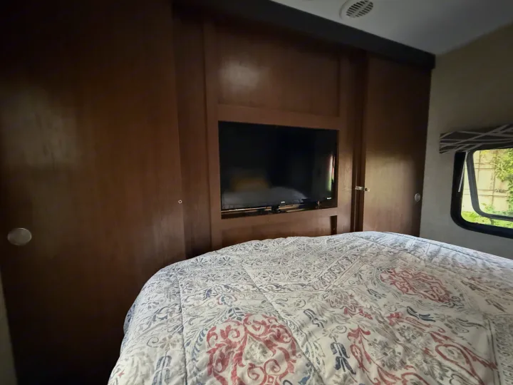 Cozy RV rental bedroom with patterned comforter, built-in TV, and wood interior. Window view shows greenery outside.. Keystone RV Hideout Luxury 2017