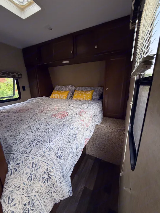 Cozy RV rental bedroom features a patterned bed, "Camping Awaits" pillows, dark wood cabinets, and large windows.. Keystone RV Hideout Luxury 2017