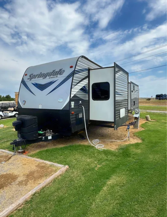 This RV rental is set up at a grassy campsite under a cloudy sky, with its slide-out extended and hookups connected.. Keystone RV Springdale 2020