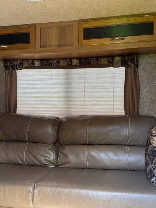 RV rental interior: A cozy brown sofa with white stitching, window with pleated blinds, and wood overhead cabinets.. Coachmen Catalina 2016
