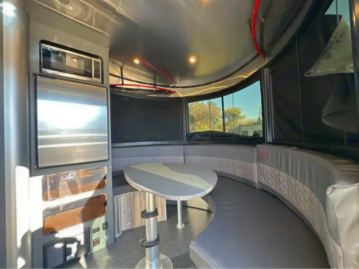 Modern RV rental interior: microwave, fridge, and curved dark seating around a table, with large scenic windows.. Airstream Basecamp 2022