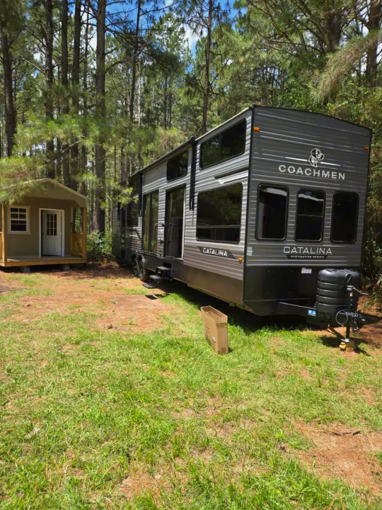 A large gray Coachmen Catalina **rv rental** is parked next to a small shed in a sunny clearing surrounded by pine trees.. Coachmen Catalina 42CONDO 2026