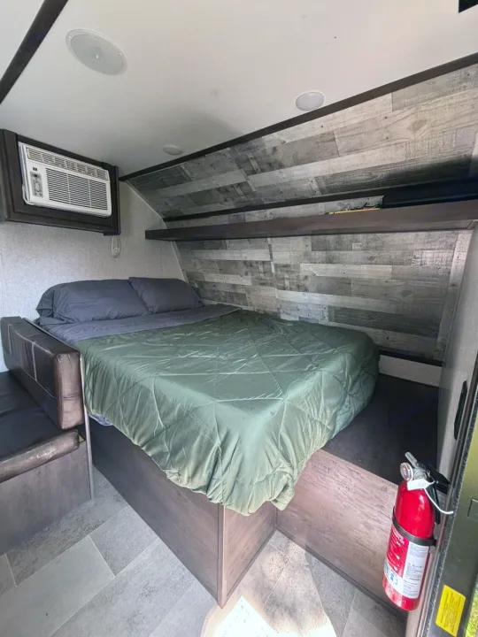 Cozy rv rental bedroom with a bed, AC unit, and shelves. A fire extinguisher is mounted near the door, featuring wood-look walls.. Dutchmen Coleman Lantern LT 2021