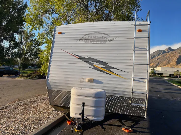 White travel trailer, possibly an RV rental, with a graphic design, front propane tanks, and a side ladder, parked outdoors with mountains & trees in background.. Eclipse Recreational Vehicles Attitude 2007