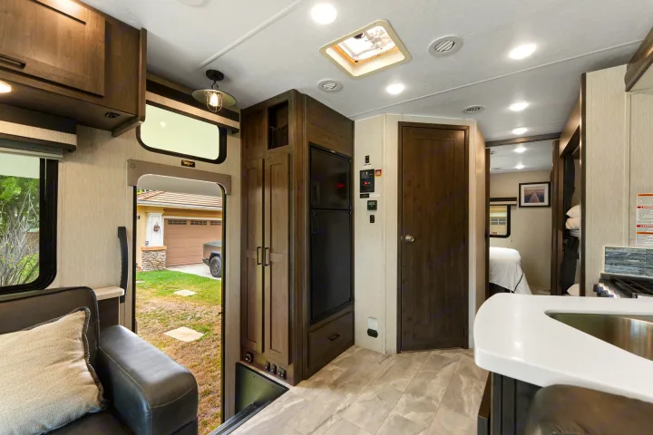 Spacious rv rental interior featuring a living area, kitchen with fridge, wood cabinets, and bedroom access. Door opens to outside.. Coachmen Pursuit 2024