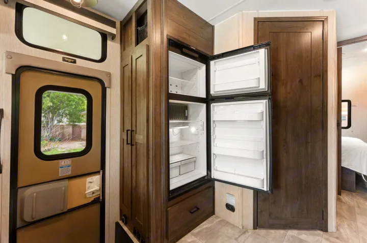 This RV rental shows an open refrigerator, adjacent dark wood cabinets, and an entry door with a window looking out to trees and a fence.. Coachmen Pursuit 2024